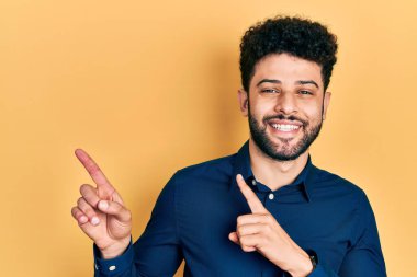 Sakallı, rahat gömlekli, gülümseyen ve kameraya bakan genç bir adam. İki eliyle ve parmaklarıyla yan tarafa bakıyor.. 