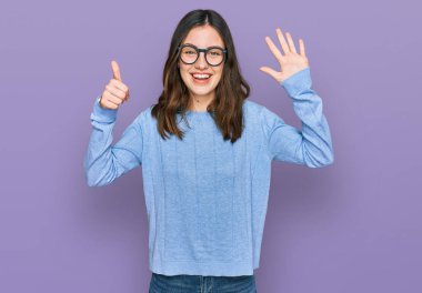 Genç ve güzel bir kadın günlük kıyafetler ve gözlük takıyor. Altıncı parmaklarıyla işaret ediyor. Kendinden emin ve mutlu bir şekilde gülümsüyor.. 
