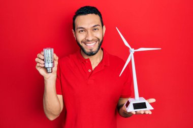 Sakallı İspanyol bir adam yenilenebilir elektrik için güneş değirmeni tutuyor ve yüzünde mutlu ve serin bir gülümsemeyle bir ampul oluşturuyor. diş gösterme. 