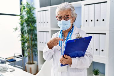 Yaşlı kadın, gri saçlı, doktor üniforması giyen ve güvenlik maskesi takan mutlu bir şekilde elini ve parmağını gösteren. 