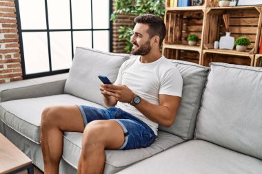 Genç İspanyol adam evde akıllı telefon kullanarak kendinden emin gülümsüyor.
