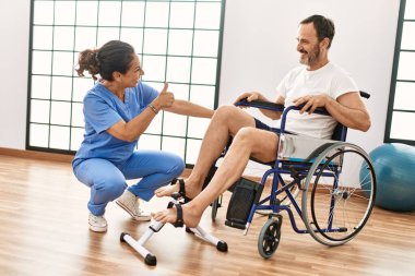 Orta yaşlı bir adam ve kadın fizik tedavi kliniğinde tekerlekli sandalyede pedal çeviriyorlar.