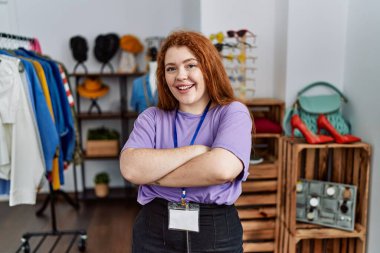 Perakende mağazasında yönetici olarak çalışan genç kızıl saçlı kadın kollarını kavuşturup kameraya bakarken gülümsüyor. pozitif kişi. 