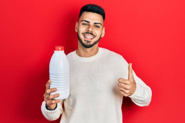 Sakallı, elinde litrelik süt şişesi olan, mutlu ve pozitif gülümseyen genç İspanyol adam, başparmağıyla mükemmel ve onay işareti yapıyor. 