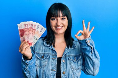 Genç İspanyol kadın elinde İzlanda Krona banknotları tutuyor tamam işareti yapıyor parmaklarıyla, gülümseyerek mükemmel bir sembol oluşturuyor. 