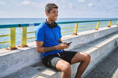 Genç Latin adam sahilde kulaklık ve akıllı telefon kullanarak mutlu gülümsüyor..