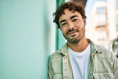 Yakışıklı, sakallı, güneşli bir günde mutlu bir şekilde gülümseyen bir İspanyol. Duvara yaslanmış.