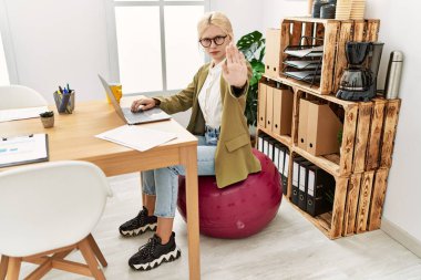 Ofiste çalışan beyaz, güzel bir iş kadını pilates topunda oturuyor ve elinde dur işareti, ciddi ve kendinden emin bir ifade, savunma jesti yapıyor. 