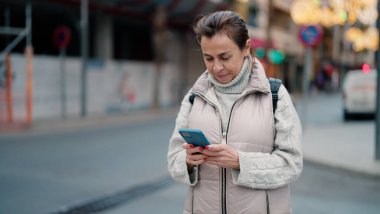 Orta yaşlı İspanyol kadın sokakta akıllı telefon kullanarak gülümsüyor.