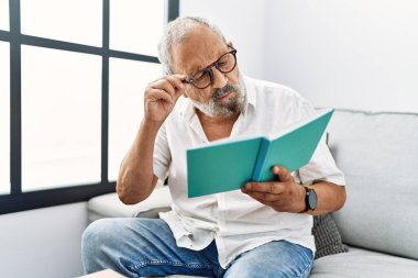 Yaşlı gri saçlı adam evde kitap okuyor.