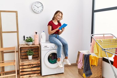 Dokunmatik ped kullanan genç beyaz bir kız evde çamaşır makinesinin başında oturmuş bekliyor..