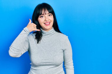 Gündelik kıyafetler giymiş, gülümseyen, el ve parmaklarıyla telefon hareketi yapan, telefonda konuşur gibi genç İspanyol bir kadın. iletişim kavramları. 