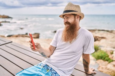 Genç kızıl saçlı turist akıllı telefon kullanıyor sahildeki bankta oturuyor..