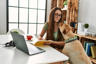 Genç İspanyol kadın dizüstü bilgisayar kullanıyor ve kitap yazıyor. Evde köpekle birlikte masada oturuyor.