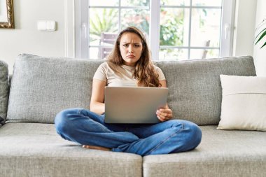Kanepede oturan genç, güzel esmer kadın evde bilgisayar laptopu kullanıyor şüpheci ve gergin, sorun yüzünden surat asıyor. negatif kişi. 