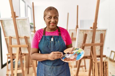 Kıdemli Afro-Amerikan kadın sanat stüdyosunda palet ve boya fırçası tutarak gülümsüyor.