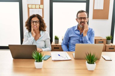 Orta yaşlı İspanyol kadın ve erkek ofiste dizüstü bilgisayarlarıyla oturup diş ağrısı ya da diş ağrısı yüzünden ağızlarına elleriyle dokunuyorlar. dişçi 