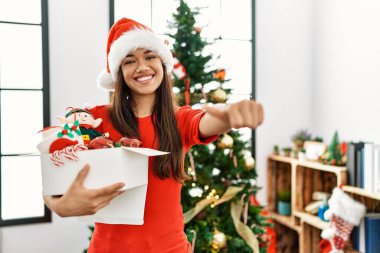 Genç esmer kadın Noel ağacının yanında duruyor elinde süslemeyle olumlu bir jest yapmayı onaylıyor, gülümsüyor ve başarı için mutlu. kazanan jest. 