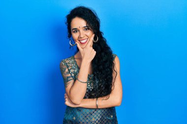 Bindi ve geleneksel kurta elbisesi giyen genç bir kadın kameraya kollarını kavuşturup gülümseyerek ve çenesini kaldırarak bakıyor. Olumlu düşünmek. 