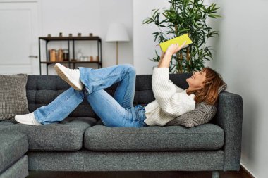 Evdeki kanepede uzanmış kitap okuyan beyaz bir kadın.