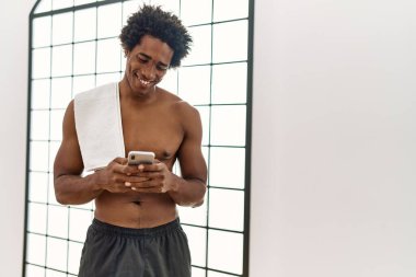 Genç Afrikalı Amerikalı adam spor salonunda akıllı telefon kullanarak mutlu bir şekilde gülümsüyor.