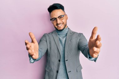 Genç Arap adam iş kıyafetleri içinde kameraya bakıp sarılmak için kollarını açmış gülümsüyor. Mutluluğu kucaklayan neşeli bir ifade.. 