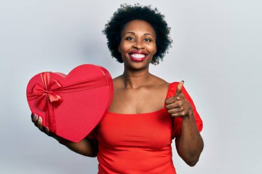 Genç Afrikalı Amerikalı kadın sevgililer günü hediyesini tutuyor mutlu ve pozitif gülümsüyor, başparmağıyla mükemmel ve onay işareti yapıyor. 