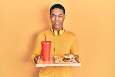 Afro-Amerikan genç adam lezzetli bir klasik burger yiyor kameraya seksi bir ifadeyle bakıyor, neşeli ve mutlu bir yüzle.. 
