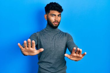 Balıkçı yaka kazak giyen sakallı bir Arap, avuçlarıyla hareket etmeyi, sinirli ve hayal kırıklığına uğramış yüz ifadesini durduruyor. 