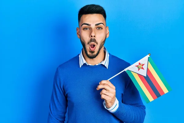 Joven Hombre Hispano Con Barba Sosteniendo Bandera Zimbabwe Asustado Sorprendido —  Fotos de Stock