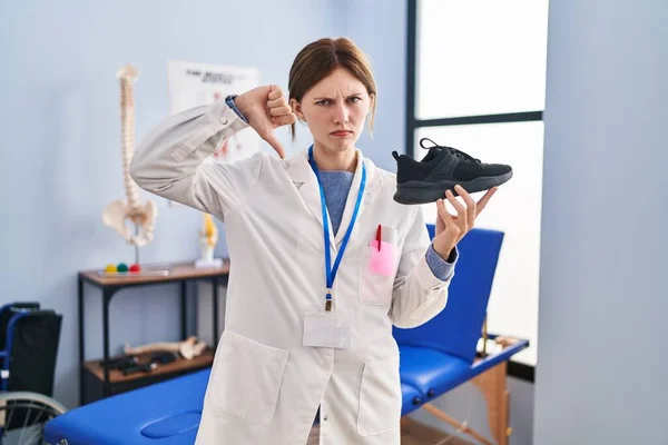 Joven Fisioterapeuta Mujer Sosteniendo Zapatillas Con Cara Enojada Signo Negativo —  Fotos de Stock