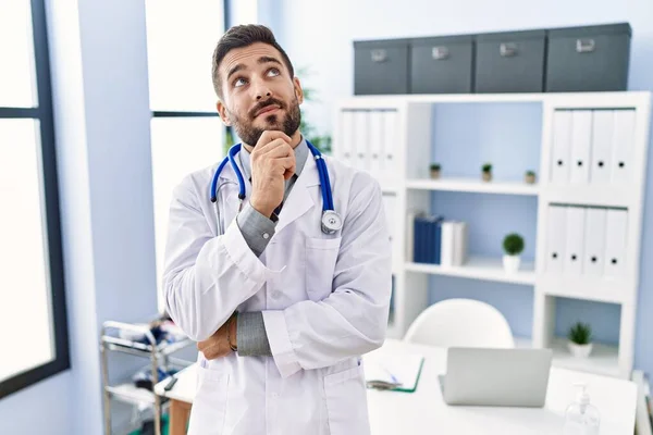 在诊所里穿着医生制服和听诊器的英俊惊慌失措的男人严肃地思考着手托着下巴的问题 思考着令人困惑的想法 — 图库照片