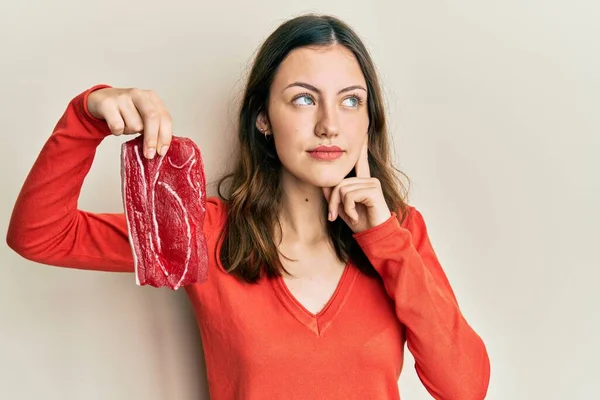 年轻的黑发女子牵着生牛肉牛排严肃的脸 手托着下巴思考着问题 思考着困惑的想法 — 图库照片