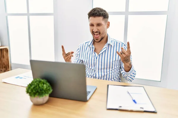 コンピューターのラップトップを使ってオフィスで働く髭を生やした若いハンサムな男が手を上げて岩のシンボルをやって狂気の表情で叫んでいます 音楽スター 重い概念は — ストック写真