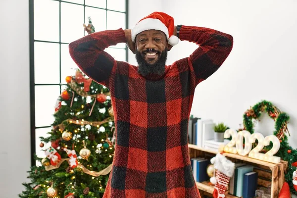 アフリカ系アメリカ人の男はサンタの帽子をかぶってクリスマスツリーのそばに立ってリラックスしてストレッチ 腕と手頭と首の後ろに笑顔幸せ — ストック写真