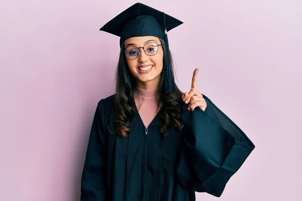 自信満々の笑顔で卒業キャップと式のローブを身に着けている若いヒスパニック系の女性が現れ 指第1位で指摘しながら — ストック写真