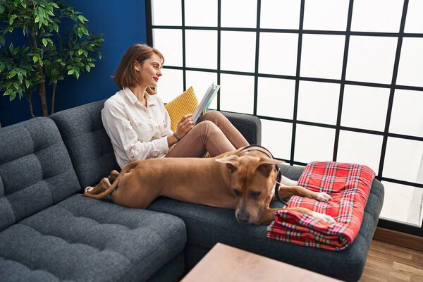 Young caucasian woman reading book sitting on sofa with dog at home