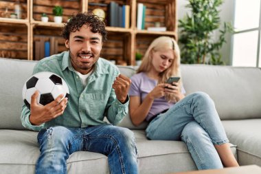 Adam mutlu bir şekilde futbol maçı izliyor ve kız arkadaşı evde akıllı telefon kullanıyor..