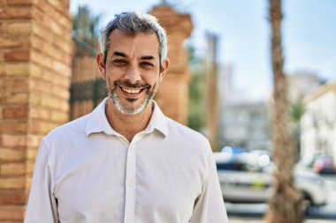 Orta yaşlı, kır saçlı, gülümseyen bir adam şehirde mutlu bir şekilde duruyor..