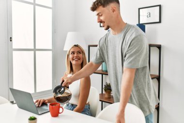 Kadın evde dizüstü bilgisayar kullanıyor ve erkek arkadaşı kahve dolduruyor..