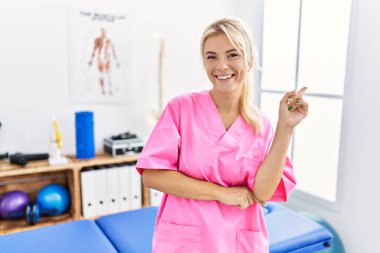 Ağrı kesici kliniğinde çalışan beyaz bir kadın yüzünde kocaman bir gülümsemeyle kameraya el ve parmak ile bakıyor.. 