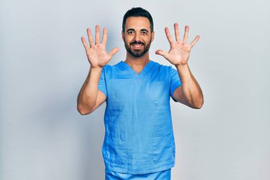 Sakallı, mavi erkek hemşire üniforması giyen yakışıklı İspanyol adam on numarayı gösterip işaret ederken kendinden emin ve mutlu gülümsüyor.. 