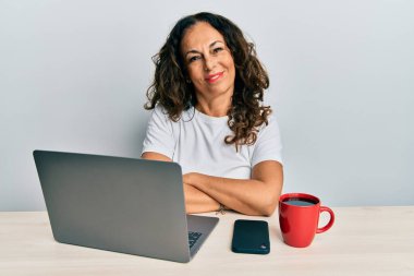 Ofiste çalışan orta yaşlı güzel bir kadın bilgisayarda çalışırken kollarını kavuşturup kameraya bakarken gülümseyen mutlu bir yüz kullanıyor. pozitif kişi. 