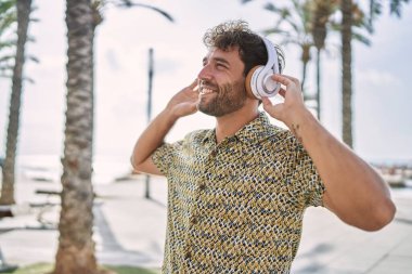 Genç İspanyol adam deniz kenarında müzik dinlerken kendinden emin bir şekilde gülümsüyor.