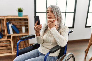 Orta yaşlı, kır saçlı, evinde tekerlekli sandalyede oturan bir kadın.
