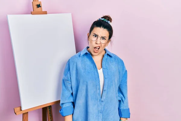 若いヒスパニック系の女性が画家のイーゼルスタンドでショックを顔に立って 懐疑的で皮肉 開いて口で驚いている — ストック写真