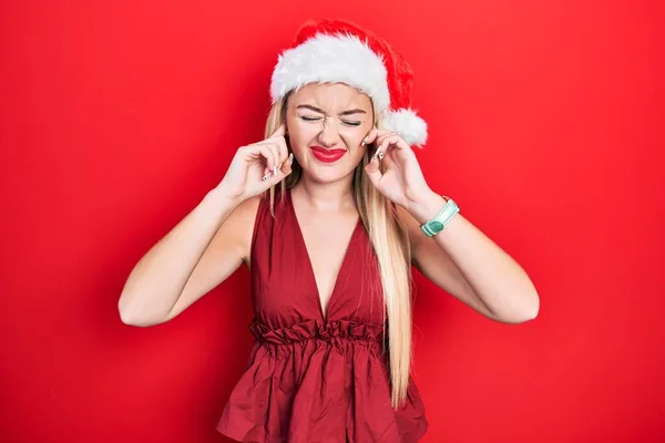 若いブロンドの女の子は 大きな音楽のノイズのためにいらいらした表情で指で耳を覆うクリスマスの帽子をかぶっています 聴覚障害の概念 — ストック写真