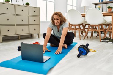 Orta yaşlı, beyaz bir kadın evinde dizüstü bilgisayar kullanarak gülümsüyor.