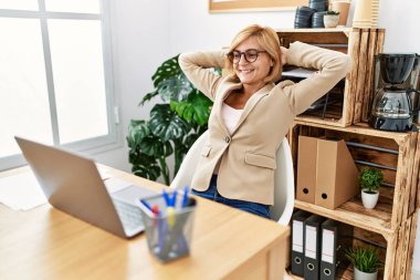 Orta yaşlı sarışın kadın, elleri ofiste rahat.