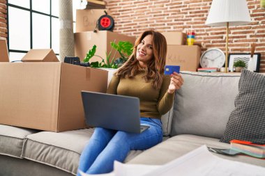 Laptop ve kredi kartı kullanan genç Latin kadın evdeki koltukta oturuyor.